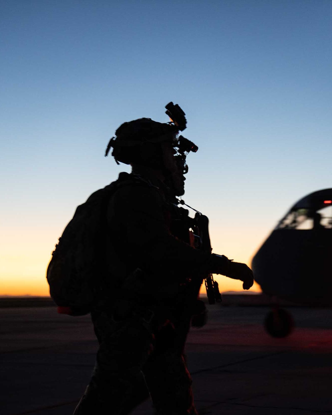 An operator in a night-vision helmet and pack silhouetted against dusk as he walks past an aircraft
